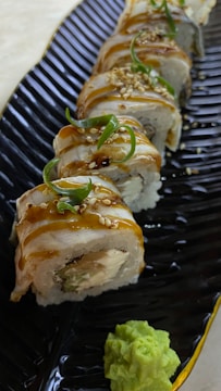 A beautifully arranged serving of sushi rolls garnished with sesame seeds and green garnish. Each roll appears to be drizzled with a sauce, and the sushi is placed on a textured dark plate. A dollop of wasabi is placed at the side.