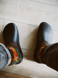 A close-up view of feet wearing dark clogs paired with colorful, patterned socks. The floor is made of light wood with visible grain textures.