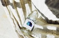 A bottle of vodka with a modern label design, placed on a textured, striped fabric. The scene is illuminated with bright, natural light, casting defined shadows and enhancing the textures.