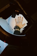 A hand wearing a watch is pressed against a photocopying machine, surrounded by papers. The watchband is dark and the device appears to be in use, as indicated by the date and time display glowing in red.