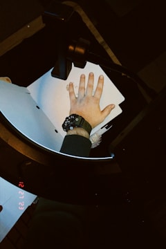 A hand wearing a watch is pressed against a photocopying machine, surrounded by papers. The watchband is dark and the device appears to be in use, as indicated by the date and time display glowing in red.