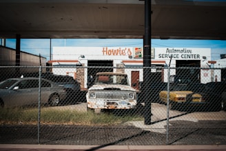 An old and rusty white truck is parked behind a chain-link fence in front of a building labeled 'Howie's Automotive Service Center.' The scene includes grass growing around the truck, a black car on its left, and a yellow car on its right. The building has signs for Castrol GTX and appears to be an automotive repair shop.