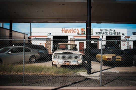 An old and rusty white truck is parked behind a chain-link fence in front of a building labeled 'Howie's Automotive Service Center.' The scene includes grass growing around the truck, a black car on its left, and a yellow car on its right. The building has signs for Castrol GTX and appears to be an automotive repair shop.