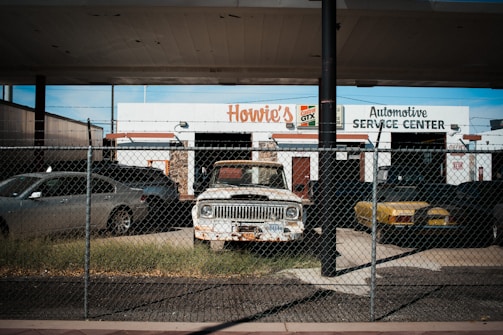 An old and rusty white truck is parked behind a chain-link fence in front of a building labeled 'Howie's Automotive Service Center.' The scene includes grass growing around the truck, a black car on its left, and a yellow car on its right. The building has signs for Castrol GTX and appears to be an automotive repair shop.
