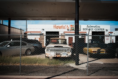 An old and rusty white truck is parked behind a chain-link fence in front of a building labeled 'Howie's Automotive Service Center.' The scene includes grass growing around the truck, a black car on its left, and a yellow car on its right. The building has signs for Castrol GTX and appears to be an automotive repair shop.