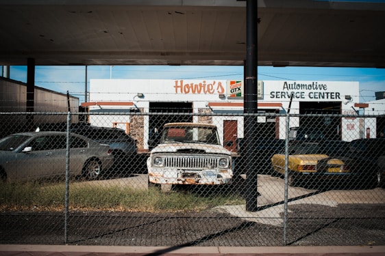 An old and rusty white truck is parked behind a chain-link fence in front of a building labeled 'Howie's Automotive Service Center.' The scene includes grass growing around the truck, a black car on its left, and a yellow car on its right. The building has signs for Castrol GTX and appears to be an automotive repair shop.
