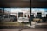 An old and rusty white truck is parked behind a chain-link fence in front of a building labeled 'Howie's Automotive Service Center.' The scene includes grass growing around the truck, a black car on its left, and a yellow car on its right. The building has signs for Castrol GTX and appears to be an automotive repair shop.