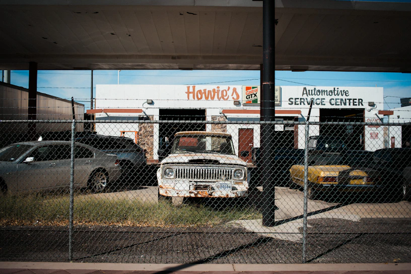 An old and rusty white truck is parked behind a chain-link fence in front of a building labeled 'Howie's Automotive Service Center.' The scene includes grass growing around the truck, a black car on its left, and a yellow car on its right. The building has signs for Castrol GTX and appears to be an automotive repair shop.