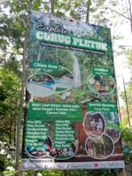 A promotional signboard for Curug Pletuk located in a lush, green area. The sign includes various offerings such as a camping package for the new year, starting December 2, 2023, to January 2, 2024. It features images of natural attractions, such as a waterfall and a pool, as well as local culinary highlights. The board details package prices and amenities like free parking and durian. Contact information and social media handles are visible at the bottom.
