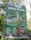 A promotional signboard for Curug Pletuk located in a lush, green area. The sign includes various offerings such as a camping package for the new year, starting December 2, 2023, to January 2, 2024. It features images of natural attractions, such as a waterfall and a pool, as well as local culinary highlights. The board details package prices and amenities like free parking and durian. Contact information and social media handles are visible at the bottom.