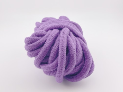 A coiled purple rope with a detailed texture, resting against a plain white background. The rope appears to be thick and tightly wound, showcasing its intricate woven pattern.