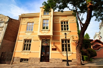 A beautifully preserved historic building with ornate architectural details, featuring cream-colored walls and large windows. The structure has intricate moldings and decorative elements around the windows and doors. In front of the building, a black streetlamp stands next to a tall, leafy tree, with other trees and bushes surrounding the area.