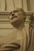A classical-style marble bust of a bearded man, draped in a toga, set against an ornate architectural background. The sculpture exhibits fine detailing and a lifelike expression, capturing the essence of historical art.