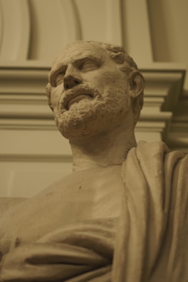 A classical-style marble bust of a bearded man, draped in a toga, set against an ornate architectural background. The sculpture exhibits fine detailing and a lifelike expression, capturing the essence of historical art.