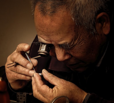 An elderly craftsman is intensely focused on working with delicate tools and a small mechanical part, possibly a watch. He is using a magnifying glass attached to his eye for detailed inspection. The setting appears to have warm lighting, highlighting the fine lines and focused expression on his face.
