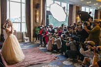 A bride in an elaborate wedding dress is posing, surrounded by a large group of photographers capturing her with professional cameras. The scene is set indoors, with natural light coming through large windows and additional lighting equipment in use.