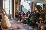 A bride in an elaborate wedding dress is posing, surrounded by a large group of photographers capturing her with professional cameras. The scene is set indoors, with natural light coming through large windows and additional lighting equipment in use.