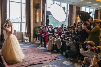 A bride in an elaborate wedding dress is posing, surrounded by a large group of photographers capturing her with professional cameras. The scene is set indoors, with natural light coming through large windows and additional lighting equipment in use.