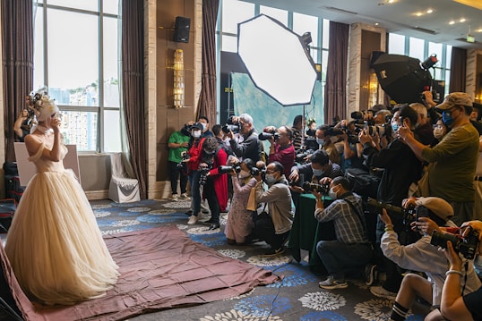 A bride in an elaborate wedding dress is posing, surrounded by a large group of photographers capturing her with professional cameras. The scene is set indoors, with natural light coming through large windows and additional lighting equipment in use.