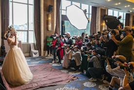 A bride in an elaborate wedding dress is posing, surrounded by a large group of photographers capturing her with professional cameras. The scene is set indoors, with natural light coming through large windows and additional lighting equipment in use.