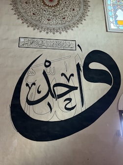 A detailed piece of Arabic calligraphy is prominently displayed on a beige wall, written in bold black strokes. Above it, an intricate decorative pattern is present, with complex geometric shapes and a vibrant central design. To the right, the edge of a stained glass window is visible, featuring colorful geometric designs.