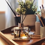 A wooden tray is placed on a table, holding a reed diffuser with black sticks in a clear glass jar containing amber-colored liquid. In the background, there is a plant with small green leaves in a white pot. To the side, several paintbrushes are standing upright in a cylindrical holder. The setting is warmly lit, creating a cozy and inviting atmosphere.
