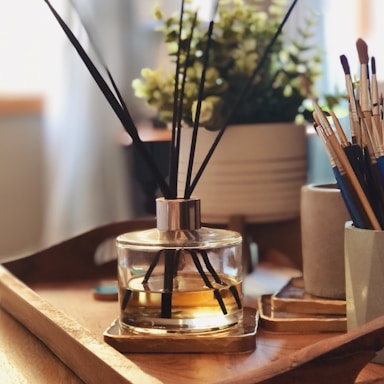 A wooden tray is placed on a table, holding a reed diffuser with black sticks in a clear glass jar containing amber-colored liquid. In the background, there is a plant with small green leaves in a white pot. To the side, several paintbrushes are standing upright in a cylindrical holder. The setting is warmly lit, creating a cozy and inviting atmosphere.