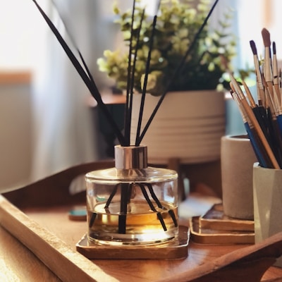 A wooden tray is placed on a table, holding a reed diffuser with black sticks in a clear glass jar containing amber-colored liquid. In the background, there is a plant with small green leaves in a white pot. To the side, several paintbrushes are standing upright in a cylindrical holder. The setting is warmly lit, creating a cozy and inviting atmosphere.