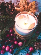 A white candle with decorative dried petals is lit and placed among pine cones, evergreen branches, cranberries, and a large yellow-brown autumn leaf. The background features a rustic texture, enhancing the warm and cozy atmosphere.