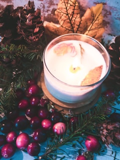 A white candle with decorative dried petals is lit and placed among pine cones, evergreen branches, cranberries, and a large yellow-brown autumn leaf. The background features a rustic texture, enhancing the warm and cozy atmosphere.