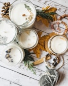 Several candles are arranged on a wooden surface, each in a glass container, with various decorations such as dried flowers, herbs, and pine cones around them. The setup includes autumn leaves and wooden beads, enhancing the rustic and cozy atmosphere.