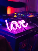A dimly lit setting features DJ equipment on a table, highlighted by a vibrant pink neon sign shaped like the word 'love.' The background is softly blurred with warm lights, suggesting a lively, energetic atmosphere commonly associated with nightclubs or parties.