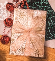 A beautifully wrapped gift box is adorned with gold snowflake patterns and a large, glittery ribbon bow. Accompanying the present are shiny red jingle bells attached to a red and white twisted string. The background features a wooden surface partially covered with decorative papers in white and dark green, each with intricate patterns.