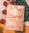 A beautifully wrapped gift box is adorned with gold snowflake patterns and a large, glittery ribbon bow. Accompanying the present are shiny red jingle bells attached to a red and white twisted string. The background features a wooden surface partially covered with decorative papers in white and dark green, each with intricate patterns.