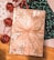 A beautifully wrapped gift box is adorned with gold snowflake patterns and a large, glittery ribbon bow. Accompanying the present are shiny red jingle bells attached to a red and white twisted string. The background features a wooden surface partially covered with decorative papers in white and dark green, each with intricate patterns.