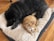 A large black dog and an orange tabby cat are lying together on a cushioned bed on a wooden floor. The dog is resting its head, while the cat is sitting alert with a collar around its neck.