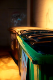 A row of industrial dumpsters with the closest one painted green, set against a dimly lit wall. Shadows and warm light create a moody atmosphere.