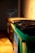 A row of industrial dumpsters with the closest one painted green, set against a dimly lit wall. Shadows and warm light create a moody atmosphere.