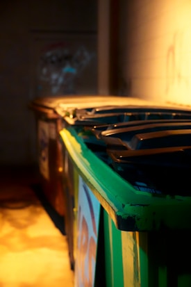 A row of industrial dumpsters with the closest one painted green, set against a dimly lit wall. Shadows and warm light create a moody atmosphere.