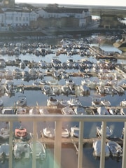 A harbor is filled with numerous small boats, docked next to each other along wooden piers. In the background, there are buildings with white facades and red roofs, typical of a seaside town. The water reflects the boats and parts of the buildings, creating a serene and tranquil atmosphere.