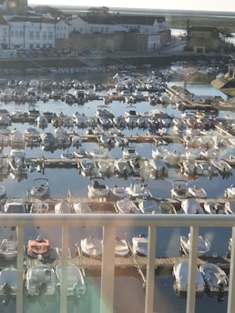 A harbor is filled with numerous small boats, docked next to each other along wooden piers. In the background, there are buildings with white facades and red roofs, typical of a seaside town. The water reflects the boats and parts of the buildings, creating a serene and tranquil atmosphere.