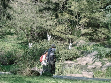 In a lush green park, two people walk along a path, surrounded by trees and shrubs. One person is carrying a backpack and the other is wearing a wide-brimmed hat. Rocks and small bushes are visible in the foreground, adding to the tranquil natural setting.