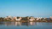 A serene lakeside view with several houses and buildings nestled among lush greenery. The calm water reflects the structures and the clear blue sky above, creating a mirror-like effect. The foreground features the lake, while the background shows a mix of residential and taller buildings.