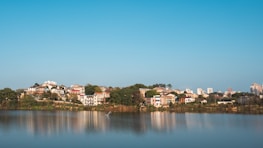 A serene lakeside view with several houses and buildings nestled among lush greenery. The calm water reflects the structures and the clear blue sky above, creating a mirror-like effect. The foreground features the lake, while the background shows a mix of residential and taller buildings.