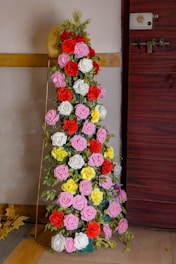 A decorative arrangement of various colored artificial flowers, including pink, red, white, and yellow roses, placed near a door. The flowers are interspersed with green leaves creating a lush display. The background includes a wall and a wooden door with a metallic lock.