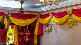 A person is decorating a room with vibrant drapery, featuring yellow and pink fabric arranged in swags. Artificial flowers in matching colors are used as embellishments. A painting decorates the wall, and soft lighting creates a warm ambiance. A hanging lamp and a ceiling fan are visible, adding to the festive atmosphere.