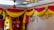 A person is decorating a room with vibrant drapery, featuring yellow and pink fabric arranged in swags. Artificial flowers in matching colors are used as embellishments. A painting decorates the wall, and soft lighting creates a warm ambiance. A hanging lamp and a ceiling fan are visible, adding to the festive atmosphere.