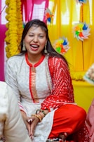 A woman dressed in traditional Indian attire, featuring a white outfit with intricate patterns and red accents, is sitting cross-legged. She is winking and smiling. The background is decorated with colorful pinwheels and yellow floral garlands, and the setting seems festive.