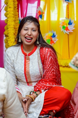 A woman dressed in traditional Indian attire, featuring a white outfit with intricate patterns and red accents, is sitting cross-legged. She is winking and smiling. The background is decorated with colorful pinwheels and yellow floral garlands, and the setting seems festive.