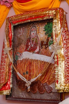 A framed artwork depicting a religious or mythological scene. At the center, a deity-like figure with a halo and ornate attire sits serenely. Below, a figure appears to be lying on a fire, possibly in a sacrificial or ritualistic manner. The background includes a swastika, trees, a horse, and additional human figures. The frame is elaborately decorated with red and gold fabric, floral designs, and a string of pearls.